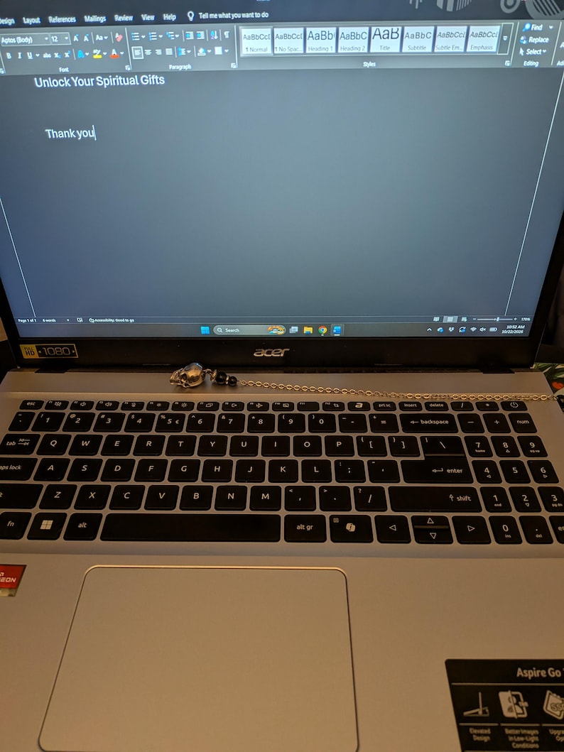May include: A silver-toned skull pendant attached to a delicate metal chain, resting on the keyboard of a silver Acer laptop. The laptop screen displays a document with the text Unlock Your Spiritual Gifts and Thank you.