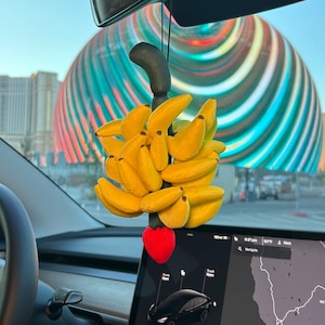 May include: A yellow banana car air freshener hanging from the rearview mirror of a car. The air freshener is shaped like a bunch of bananas with a red heart at the bottom. The car is driving past a large, colorful, striped, circular structure.