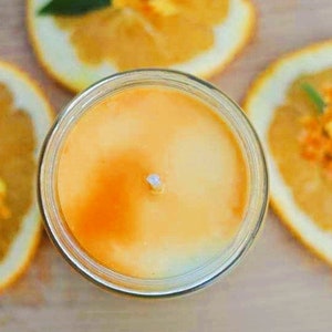 May include: A jar candle with an orange-yellow wax, surrounded by orange slices decorated with small orange flowers and green leaves. The candle has a white wick and is set on a light brown surface.