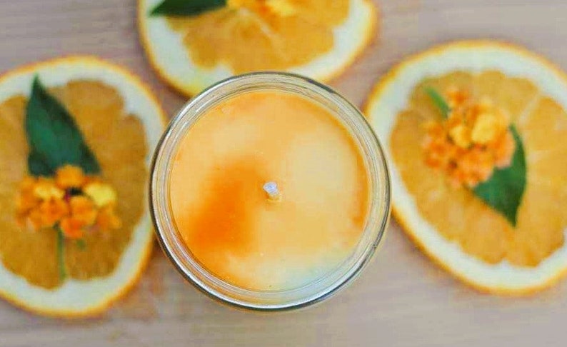May include: A jar candle with an orange-yellow wax, surrounded by orange slices decorated with small orange flowers and green leaves. The candle has a white wick and is set on a light brown surface.