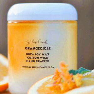 May include: A cylindrical candle with a white lid and an orange and white gradient. The label reads "Bartley's Candles ORANGECICLE 100% SOY WAX COTTON WICK HAND CRAFTED". Slices of orange are in the foreground.