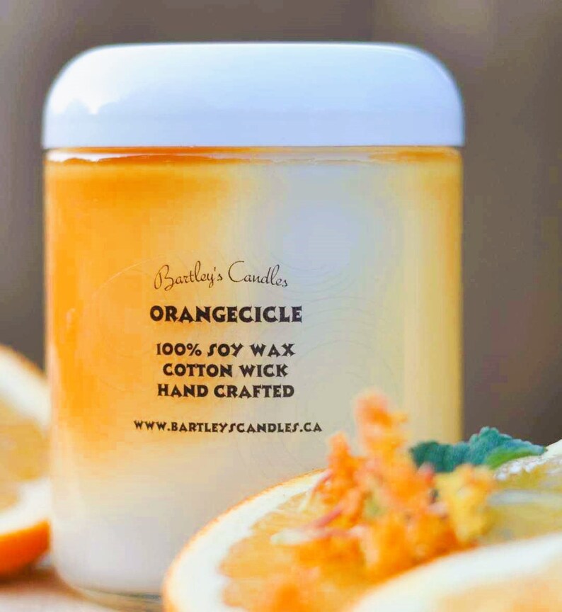 May include: A cylindrical candle with a white lid and an orange and white gradient. The label reads "Bartley's Candles ORANGECICLE 100% SOY WAX COTTON WICK HAND CRAFTED". Slices of orange are in the foreground.