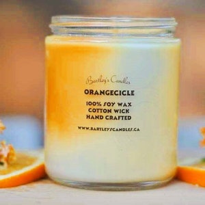 May include: A glass jar candle with an orange and white gradient. The label reads "Orangecicle" and lists "100% Soy Wax, Cotton Wick, Hand Crafted." The candle is surrounded by orange slices and orange flowers.