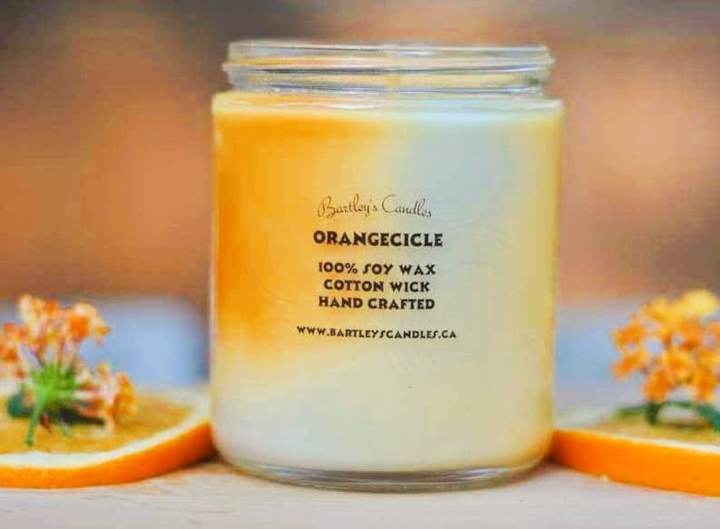 May include: A glass jar candle with an orange and white gradient. The label reads "Orangecicle" and lists "100% Soy Wax, Cotton Wick, Hand Crafted." The candle is surrounded by orange slices and orange flowers.