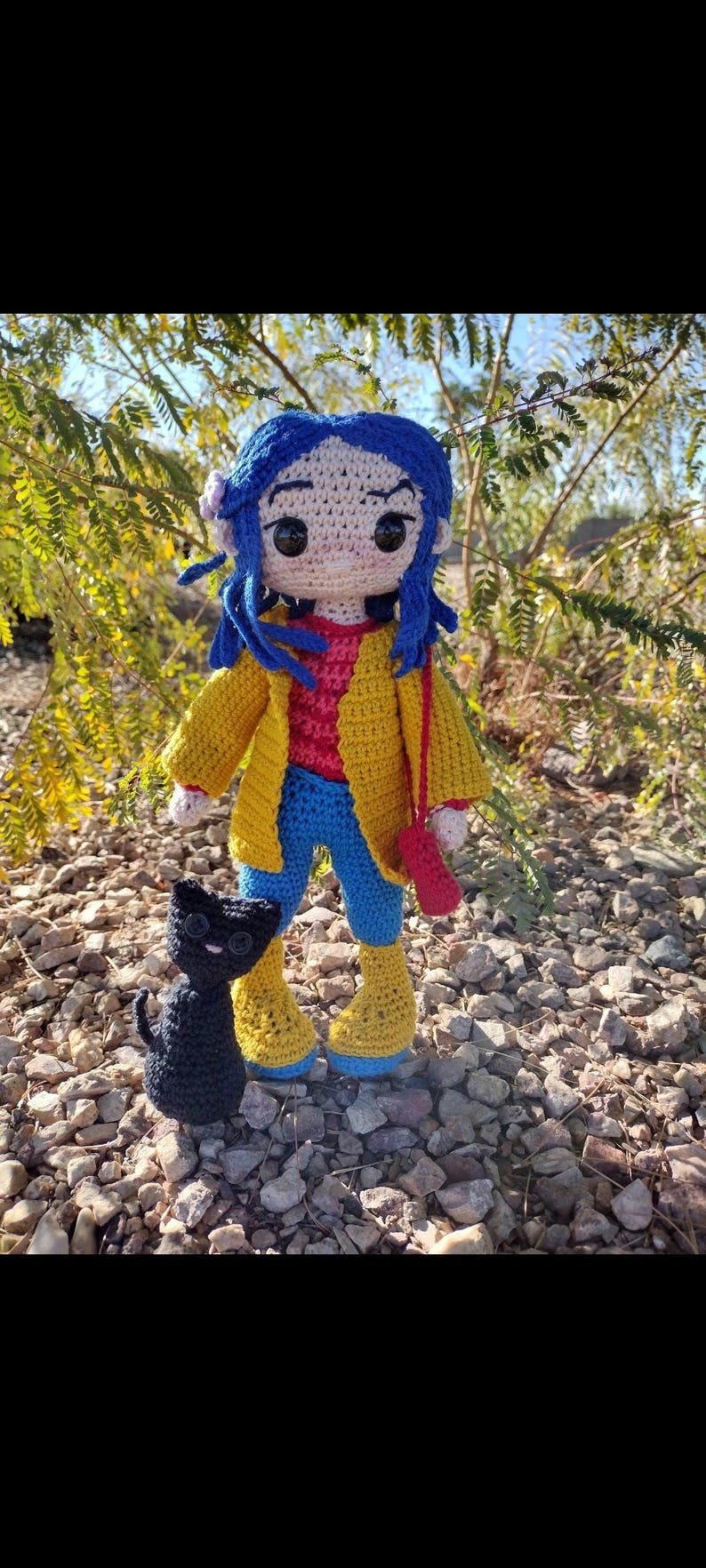 Puede incluir: Una mu&ntilde;eca de crochet con cabello azul, vistiendo una chaqueta amarilla, pantalones azules y botas amarillas. La mu&ntilde;eca lleva un bolso rojo y est&aacute; de pie junto a un gato negro de crochet.