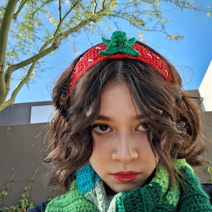 Puede incluir: Primer plano de una persona que lleva una diadema de fresa de ganchillo roja y verde. La diadema tiene una hoja verde en la parte superior y costuras blancas alrededor de los bordes. La persona lleva una bufanda verde y azul.