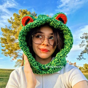May include: A green crochet frog hood with red and black accents. The hood is worn by a person wearing glasses and a white shirt. The hood has large, round eyes and is made of a textured yarn. The text "@littleguppyshop" is visible.