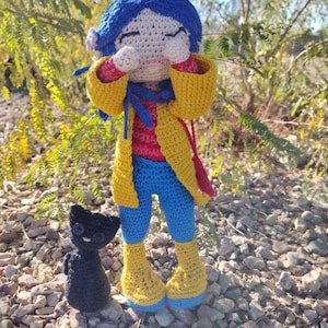 Puede incluir: Una mu&ntilde;eca de crochet con cabello azul, que lleva una chaqueta amarilla, pantalones azules y botas amarillas. La mu&ntilde;eca se est&aacute; tapando la cara con las manos y est&aacute; de pie sobre un lecho de piedras peque&ntilde;as. Un peque&ntilde;o gato negro de crochet est&aacute; de pie junto a la mu&ntilde;eca.