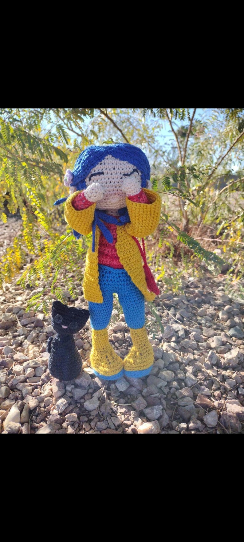 Puede incluir: Una mu&ntilde;eca de crochet con cabello azul, que lleva una chaqueta amarilla, pantalones azules y botas amarillas. La mu&ntilde;eca se est&aacute; tapando la cara con las manos y est&aacute; de pie sobre un lecho de piedras peque&ntilde;as. Un peque&ntilde;o gato negro de crochet est&aacute; de pie junto a la mu&ntilde;eca.