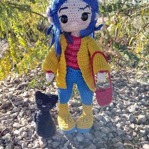 Puede incluir: Mu&ntilde;eca de crochet con cabello azul, vistiendo una chaqueta amarilla, jeans azules y botas amarillas. La mu&ntilde;eca lleva un bolso rojo de crochet. Un gato negro de crochet est&aacute; sentado frente a la mu&ntilde;eca.
