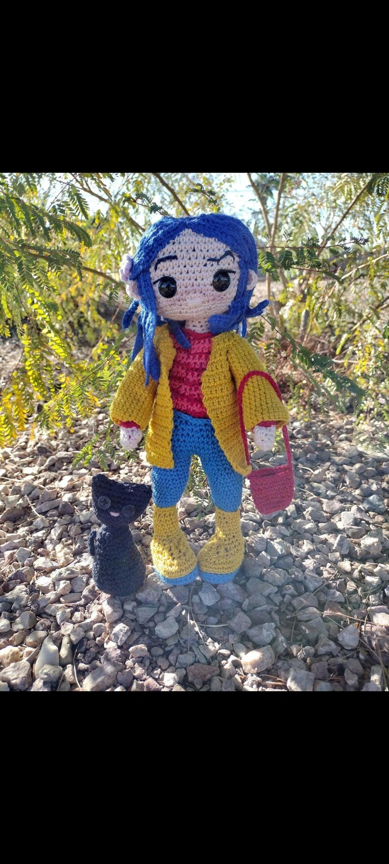Puede incluir: Mu&ntilde;eca de crochet con cabello azul, vistiendo una chaqueta amarilla, jeans azules y botas amarillas. La mu&ntilde;eca lleva un bolso rojo de crochet. Un gato negro de crochet est&aacute; sentado frente a la mu&ntilde;eca.
