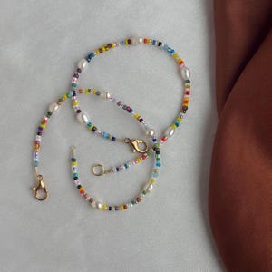 May include: A colorful beaded necklace with white pearls and gold clasps. The necklace is made of small, colorful beads and has a delicate, feminine look.