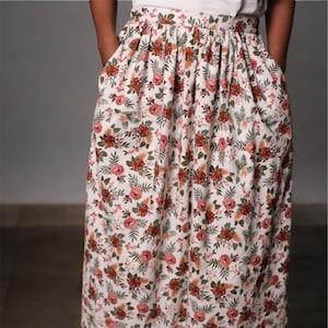 以下が含まれることがあります： 白地を背景に、赤、ピンク、緑の花柄が施されたロング丈のフローラルスカート。 スカートにはポケットがあり、白いトップスと合わせています。 スカートは足首丈です。