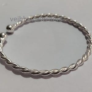 Peut inclure: Un bracelet jonc argenté avec un motif torsadé en forme de corde. Le bracelet a une finition polie et une petite boule au niveau du fermoir. Le bijou est présenté sur une surface claire.