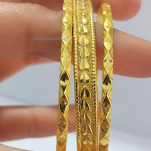 Könnte beinhalten: Drei goldene Armreifen mit aufwendigen Designs. Ein Armreif hat ein sich wiederholendes Herzmuster, während die anderen beiden ein rautenförmiges Muster aufweisen. Der Schmuck wird vor einem neutralen Hintergrund präsentiert.