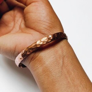 May include: A copper-colored cuff bracelet with a hammered texture. The bracelet is worn on a wrist, showcasing its warm, metallic hue and unique surface design. The cuff is a simple, yet elegant piece of jewelry.