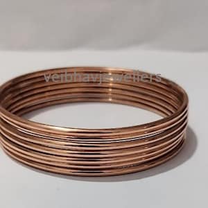May include: A stack of copper-colored bangles. The jewelry is stacked together, with the text "veirbhavjewellers" visible on the surface. The bangles are a warm, metallic color, and the image is taken against a white background.