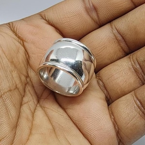 May include: A polished silver ring with a rounded, barrel-like shape. The ring has a smooth, reflective surface and a thick band. The design is simple and modern, suitable for everyday wear. The ring is shown on a hand.