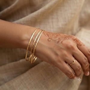 May include: Three thin, gold-toned metal bangles with a hammered, textured surface worn on the wrist, accompanied by a single, delicate gold-toned ring featuring a V-shaped chevron design worn on the ring finger of the same hand.