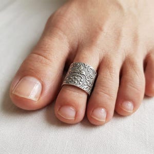 Könnte beinhalten: Ein silberfarbener Zehenring mit einem verzierten Blumen- und Scroll-Design. Der Ring ist breit und umschließt den Zeh. Der Ring wird an einem Fuß mit weißem Hintergrund getragen.