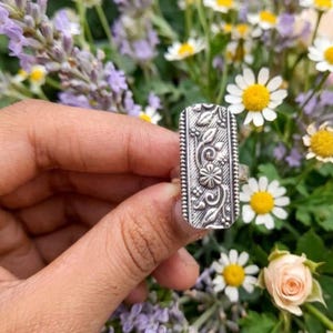 May include: A silver rectangular ring with an intricate floral pattern. The ring showcases a central flower and curling vines, enclosed by a beaded edge. The ring is held against a backdrop of flowers.