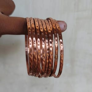 May include: A collection of copper bangles with a hammered texture. The bangles are a warm, reddish-brown color and appear to be stacked together. The hammered design adds a unique, handcrafted look to the jewelry.