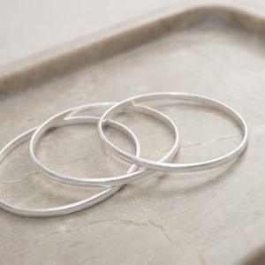 May include: Three silver-colored bangles are intertwined on a light-colored, textured surface. The bangles are circular and appear to be made of metal. The image is well-lit, highlighting the smooth texture of the jewelry.
