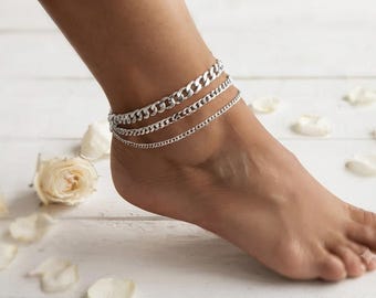 May include: A set of three silver-toned anklets worn on the ankle. The jewelry consists of one thick curb chain, one medium-sized curb chain, and one thin cable chain, all featuring a polished metallic finish and a secure clasp mechanism.