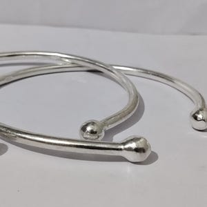 May include: Two silver-colored, open-ended bangles with a smooth, rounded ball at each end. The bracelets are simple in design and appear to be made of a shiny metal. The bangles are resting on a white surface.