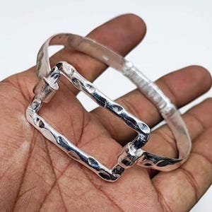 May include: A silver bracelet with a hammered texture and a rectangular clasp. The bracelet is made of a shiny metal and has a unique, handcrafted appearance. The design features a textured surface, adding visual interest to the jewelry.
