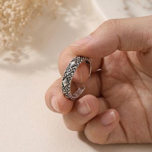 May include: An open-style silver-toned ring featuring a textured, oxidized finish with a repeating pattern of raised diamond shapes and small, granular details along the outer band, designed with a gap for adjustable sizing and a metallic sheen.