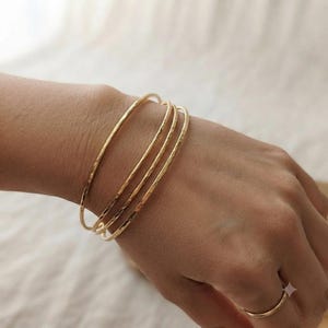 May include: A set of three thin, gold-colored metal bangles with a textured, hammered surface finish. The bracelets are worn together on the wrist, and a matching thin, plain gold-colored metal ring is worn on the finger of the same hand.