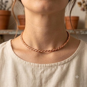 May include: A copper-colored metal choker necklace featuring a twisted rope-like design. The necklace sits closely around the neck and is constructed from a single continuous piece of metallic material with a polished, reflective finish.