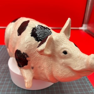 Może przedstawiać: Ceramiczna figurka świnki w kolorze białym z czarnymi plamkami. Świnka leży na boku, głowa zwrócona w prawo. Świnka ma szczegółową twarz z małym ryjkiem i czarnymi oczami.