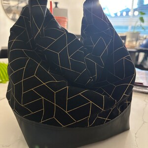 Könnte beinhalten: Eine schwarze Lunch-Tasche mit einem goldenen geometrischen Muster und einem schwarzen Lederboden. Die Tasche hat einen schwarzen Riemen mit einer Schnalle.