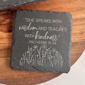 May include: Two square slate coasters with the text "SHE SPEAKS WITH wisdom AND TEACHES WITH kindness." and "PROVERBS 31:26" etched in white. A floral design is etched below the text. The coasters are on a wooden surface.