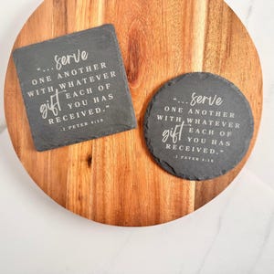 May include: Two dark gray slate coasters, one square and one circular, both featuring the engraved white text: "...serve ONE ANOTHER WITH WHATEVER EACH OF YOU HAS RECEIVED." - 1 PETER 4:10. The coasters have natural, rough-hewn edges.