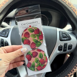 May include: Two circular car coasters packaged in a clear plastic sleeve with a white header card labeled Car Coasters Handmade with love. The coasters feature a repeating pattern of red raspberries and green leaves on a light pink background.