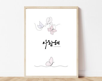 May include: A framed art print featuring delicate line drawings of butterflies in shades of pink and lavender. The Korean phrase "사랑해" (Saranghae), meaning "I love you," is written in elegant calligraphy below the butterflies.