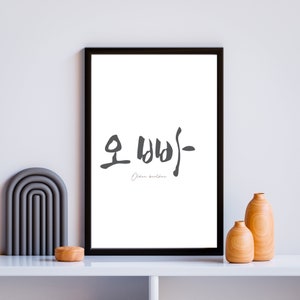 May include: Black and white framed print with the Korean word "Older Brother" written in calligraphy. The print is hanging on a white wall with a gray arch and two wooden vases.