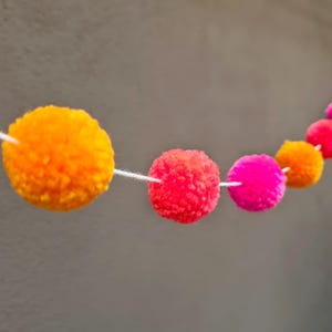 Könnte beinhalten: Eine farbenfrohe Pompon-Girlande mit orangefarbenen, roten und pinkfarbenen Pompons. Die Pompons sind mit weißem Faden aufgereiht. Diese dekorative Girlande eignet sich für festliche Anlässe.