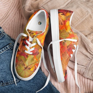 May include: A pair of white canvas low-top sneakers with a colorful autumn leaf pattern. The shoes have white laces and are laid out on a pink and blue fabric background.