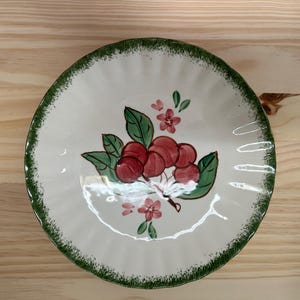 May include: A round ceramic plate with a scalloped edge featuring a green sponge-painted rim. The center of the white plate displays a hand-painted illustration of a cluster of red cherries with green leaves and small pink blossoms.
