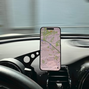 Peut inclure: Un smartphone affiche une application de cartographie, monté sur le tableau de bord d'une voiture. Le téléphone est maintenu par un support noir, près de la grille d'aération. Le volant et le tableau de bord sont visibles, avec un arrière-plan flou.