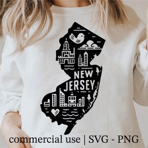 Op de afbeelding: Witte sweatshirt met een zwarte afbeelding van de staat New Jersey. Het ontwerp bevat illustraties van gebouwen, een vogel en de tekst "NEW JERSEY". De onderkant van de afbeelding luidt "commercial use | SVG - PNG".