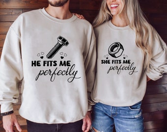 May include: Matching beige sweatshirts with black graphics. One sweatshirt features a bolt design with the text "He fits me perfectly." The other sweatshirt has a nut design with the text "She fits me perfectly."