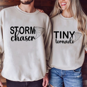 Puede incluir: Dos sudaderas beige con texto negro y un gráfico de tornado. Una dice "STORM chaser" y la otra dice "TINY tornado". Las sudaderas son un conjunto a juego.