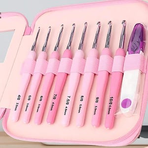 Op de afbeelding: Een roze haaknaaldset in een etui met ritssluiting. De set bevat negen haaknaalden in verschillende roze tinten, met maten variërend van 2,5 mm tot 6,0 mm. De etui is open en onthult de haken netjes gerangschikt in individuele sleuven. De tekst "Sew Need It Crafts" is zichtbaar.