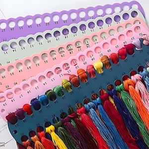 Könnte beinhalten: Eine Sammlung farbenfroher Stickgarne, die auf mehreren Kunststoff-Garnhaltern angeordnet sind. Die Halter sind in Lila-, Grün-, Rosa- und Türkistönen gehalten und haben Löcher für den Faden und nummerierte Etiketten. Die Garnfarben umfassen Rot, Blau, Grün, Gelb und Orange.