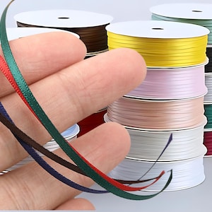 Puede incluir: Una selección de carretes de cinta de raso en varios colores, incluyendo amarillo, rosa y verde. Varios hilos de cinta de diferentes colores se sostienen frente a los carretes. Los carretes están apilados y la cinta es lisa y brillante.